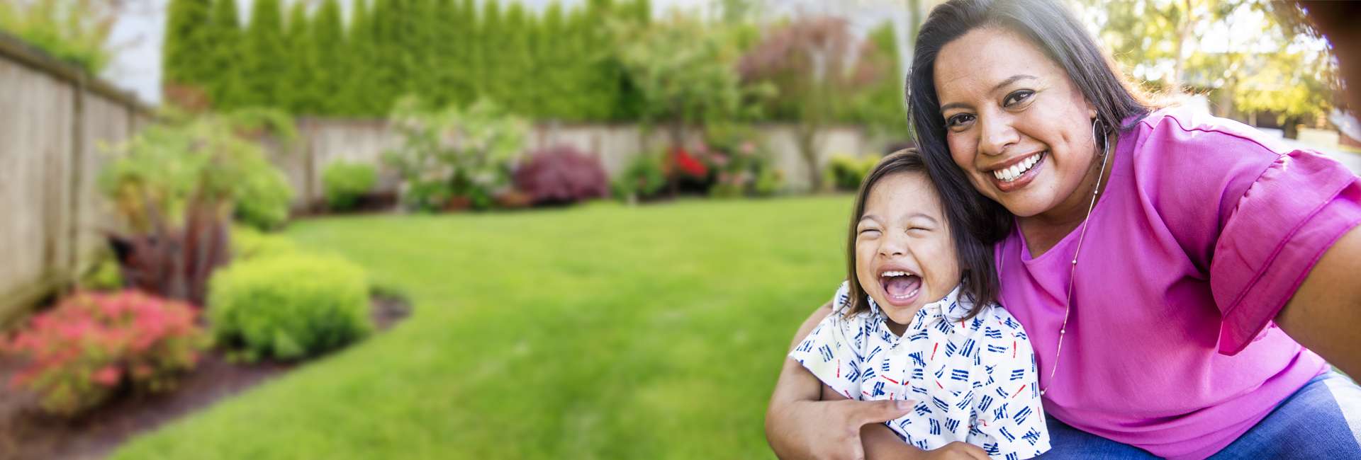 woman in a pink shirt smiling holding her laughing child in their back yard.
